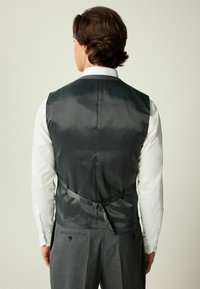 Gilet noir en satin avec une texture lisse, sangle réglable au dos, associé à une chemise blanche à boutons et un pantalon gris.