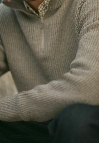 Vaaleanharmaa neulepusero, jossa on korkea kaulus ja puolivälin vetoketju. Varustettu teksturoituneella kuviolla ja istuvilla hihoilla.