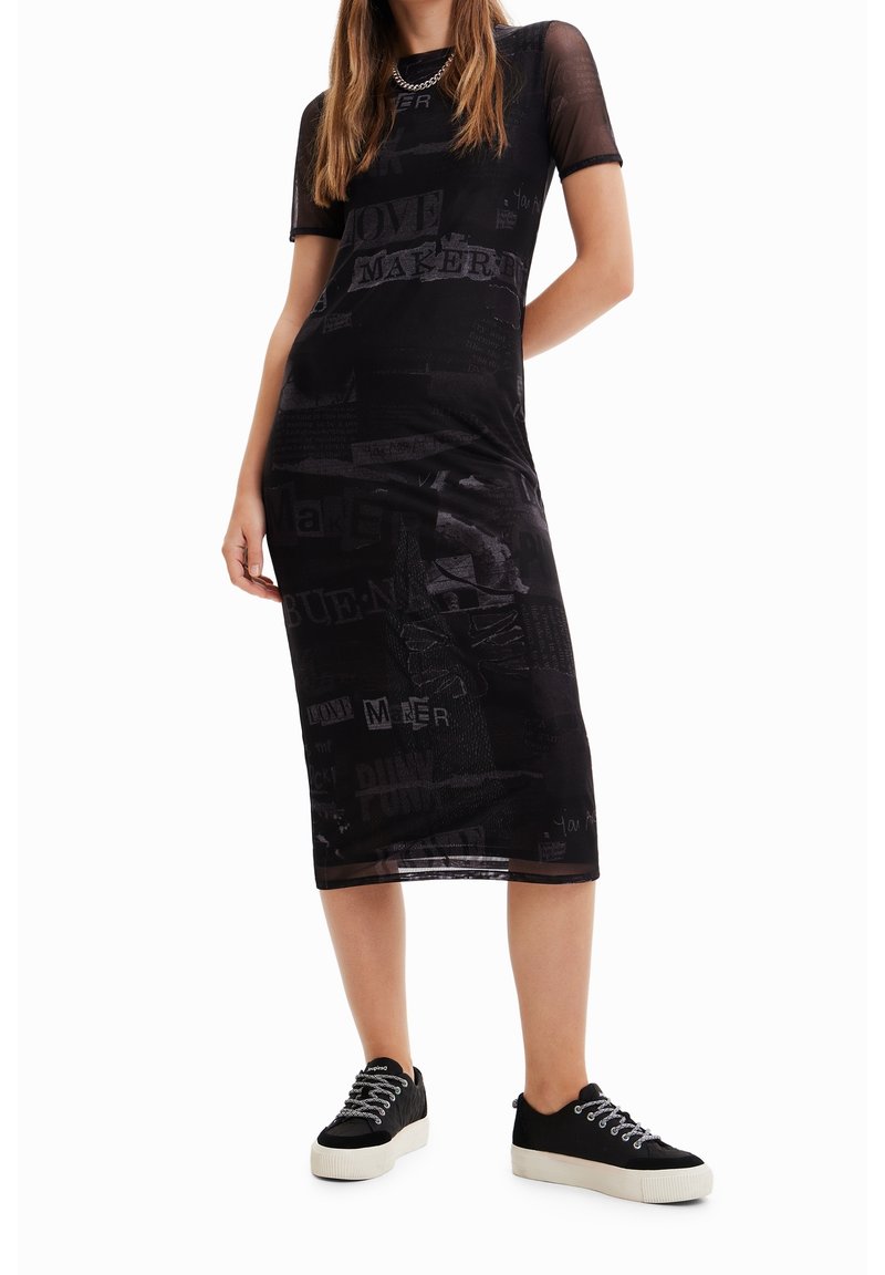 Femme portant une robe midi en maille noire avec du texte imprimé et des baskets noires avec des semelles et des lacets blancs, se tenant devant un fond blanc.