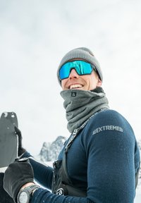 Skiausrüstung mit einem navy blauen Oberteil, grauem Schal und grauer Mütze. Enthält blaue verspiegelte Skibrillen und einen schwarzen Handschuh, mit Schnee auf der Hand.