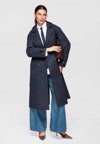 Femme avec des lunettes portant un trench bleu marine, une chemise blanche, une cravate noire, un jean à jambes larges, des talons marron, tenant un sac à main marron sur le bras.