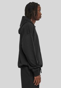 Sudadera negra hecha de un tejido suave con un ajuste holgado, bolsillo tipo canguro y cordones ajustables. Puños y dobladillo de canalé. Vista de perfil lateral.
