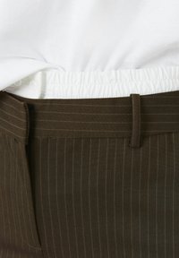 Pantalons marron à rayures fines avec une taille plate, une ceinture élastique et un tissu intérieur blanc contrastant qui ressort en haut.