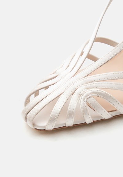 Λευκό σανδάλι με λουράκια, με πολλά λεπτά δερμάτινα λουράκια να διασταυρώνονται πάνω από τα ανοιχτά δάχτυλα σε μπεζ σόλα, πάνω σε λευκό φόντο.