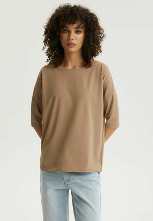 Kobieta o kręconych włosach, ubrana w luźną brązową bluzkę z półdługimi rękawami i jasnoniebieskie dżinsy, stojąca z rękami za plecami na białym tle.