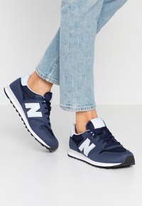 Marinblå sneakers i mesh och mocka, med en ljusblå accent på tungan och hälen, samt en vit logo och gummisula.