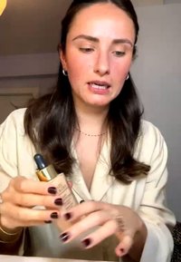 Frau mit dunklem Haar und lackierten Nägeln hält eine Foundation-Flasche mit Pipette, um deren Anwendung drinnen zu demonstrieren oder zu erklären.