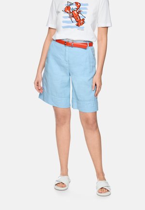 Frau trägt ein weißes T-Shirt mit rotem Hummerdruck, hellblaue knielange Shorts, roten Gürtel und weiße geflochtene Sandalen.