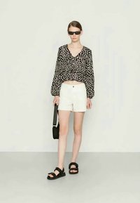Blusa de bolinhas em preto e branco com mangas bufantes, calções brancos, sandálias pretas e uma carteira preta. Modelo usa óculos de sol pretos.