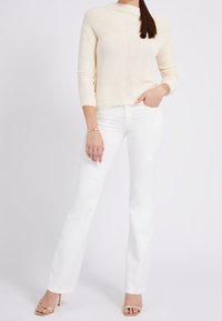 Maglione lavorato a maglia color crema con un collo alto, abbinato a jeans bianchi a zampa d'elefante e sandali con tacco dorato. Texture liscia e design aderente.