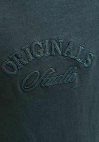 Camisa de algodón de manga corta en verde oscuro con texto bordado en relieve "ORIGINALS Studio" en un tono contrastante, con una textura suave.