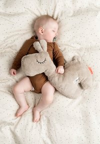 Een beige knuffelslak ligt naast een slapende baby in een bruin rompertje op een licht gevlekt deken. Het speelgoed heeft een gestikte glimlach en een zachte textuur.