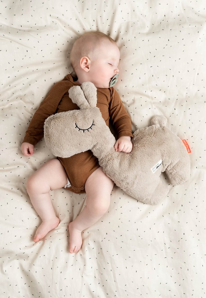 Een beige knuffelslak ligt naast een slapende baby in een bruin rompertje op een licht gevlekt deken. Het speelgoed heeft een gestikte glimlach en een zachte textuur.