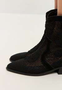 Botines negros hasta el tobillo con punta afilada, que presentan texturas de malla y terciopelo, patrones florales y un tacón bajo apilado.