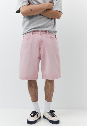 Persoon die met de armen over elkaar staat, gekleed in een grijs T-shirt, roze knielange korte broek, witte sokken en crèmekleurige sneakers met zwarte zolen.