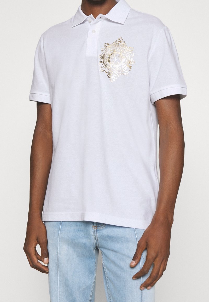 Weißes Polo-Shirt mit einem gestickten goldenen Dekor-Symbol auf der linken Brust. Kurzärmlig mit einem klassischen Kragen und weichem Stoff. 