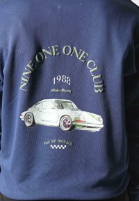 Felpa blu navy con una grafica di una macchina vintage bianca e verde, con il testo "NINE ONE ONE CLUB 1988 Atelier Racing" sotto.