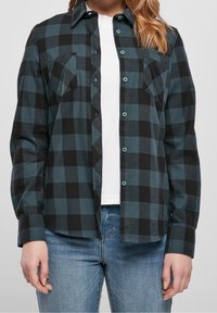 Camicia in flanella a quadri in verde scuro e nero; presenta una chiusura a bottoni, due tasche sul petto e un orlo leggermente curvo. Tessuto in cotone, maniche lunghe.