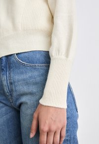 Cremefarbener, gestrickter Pullover mit gerippten Bündchen, getragen zu hellblauen, hochgeschnittenen Jeans. Der Pullover hat ein strukturiertes Maschenmuster.