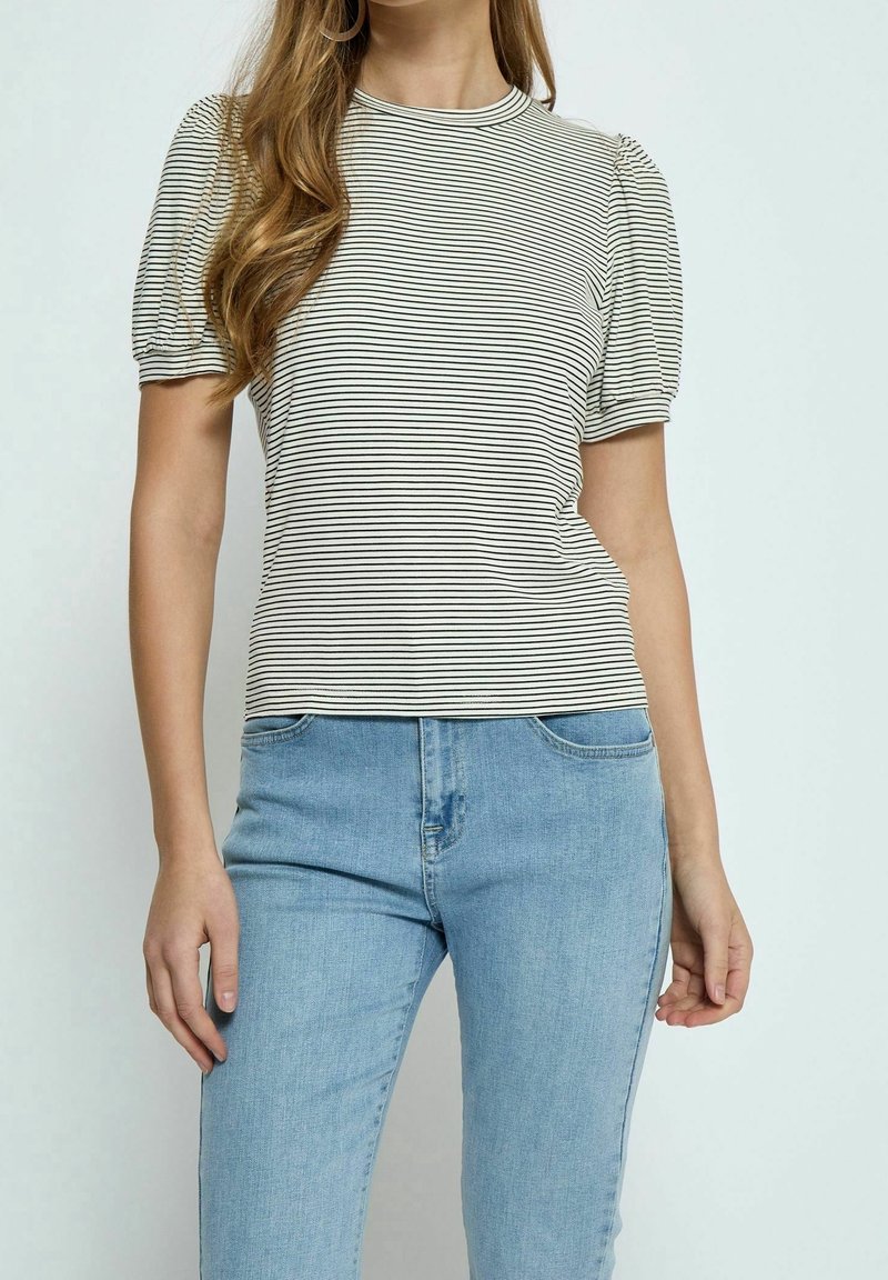 Femme portant un haut à manches bouffantes rayé blanc et noir, associé à un jean taille haute bleu clair, debout devant un fond uni.