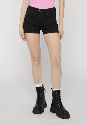 Donna che indossa pantaloncini in denim neri, calzini bianchi alla caviglia e stivali da combattimento neri con lacci, in piedi contro uno sfondo chiaro uniforme.