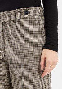 Frau trägt beige und schwarze karierte Hosen mit einem schwarzen Knopf, einen schwarzen Langarm-Top, die Hand entspannt an der Seite.