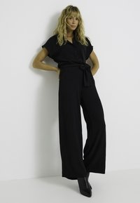 Mono negro con mangas cortas, frente abotonado y cintura anudada. Diseño de pierna ancha. Llevado con botas de tacón negro brillantes. Fondo minimalista.