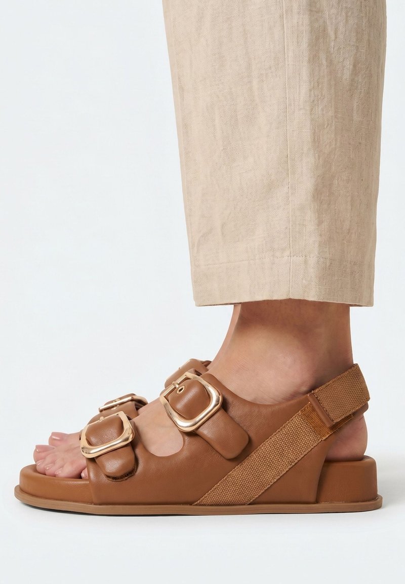 Close-up van een voet met beige cropped broek en een bruine sandaal met twee grote gouden gespen en een achterriem.