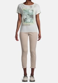 Leichtes, durchscheinendes weißes T-Shirt mit floralem Grafikdruck, kurzen Ärmeln und rundem Ausschnitt. Kombiniert mit beigen, knöchellangen Hosen und weißen Schuhen.