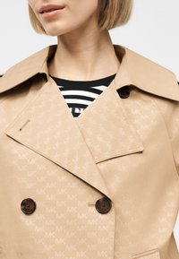 Γυναίκα που φοράει μπεζ διπλόμπουκο καμπαρντίνα με διακριτικό μοτίβο με το μονόγραμμα MK και πουκάμισο σε μαύρες και λευκές ρίγες από κάτω.