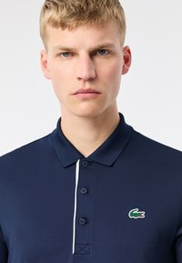 Marinblå polo tröja i texturerat tyg, med traditionell krage, knäppning fram och en liten grön krokodil-logotyp på bröstet.