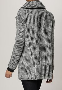 Chaqueta de punto en blanco y negro con un patrón texturizado, collar ancho y puños negros. La vista trasera muestra los detalles de las costuras y un corte generoso.