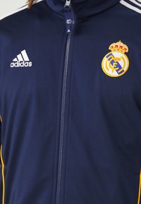 Veste zippée bleu marine en tissu léger ; doté de rayures blanches, d'un logo d'équipe brodé et de subtils accents jaunes sur les côtés.