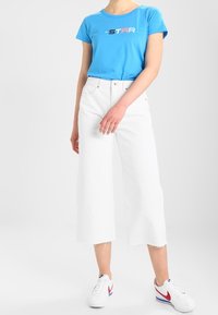 Světle modré tričko s krátkým rukávem a potiskem "ALL-STAR", v kombinaci s bílými širokými džíny a bílými teniskami s červenými akcenty.