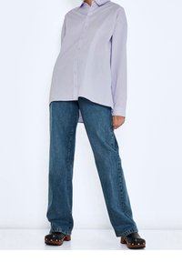 Camicia a bottoni viola chiaro con maniche lunghe, abbinata a jeans a gamba larga blu e zoccoli neri con borchie argentate sulla punta.
