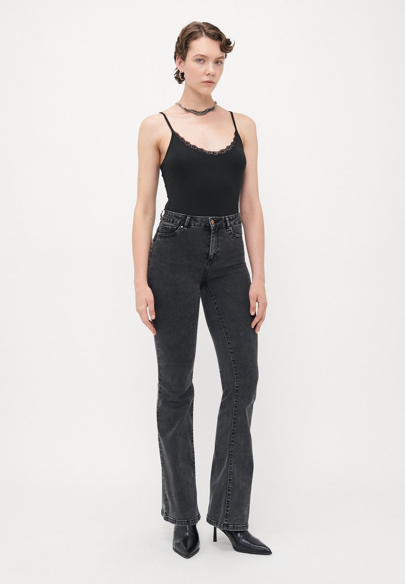 Femme portant une camisole noire avec des bordures en dentelle, un jean noir délavé taille haute, des bottes à talons noires et un collier en chaîne argentée, debout.