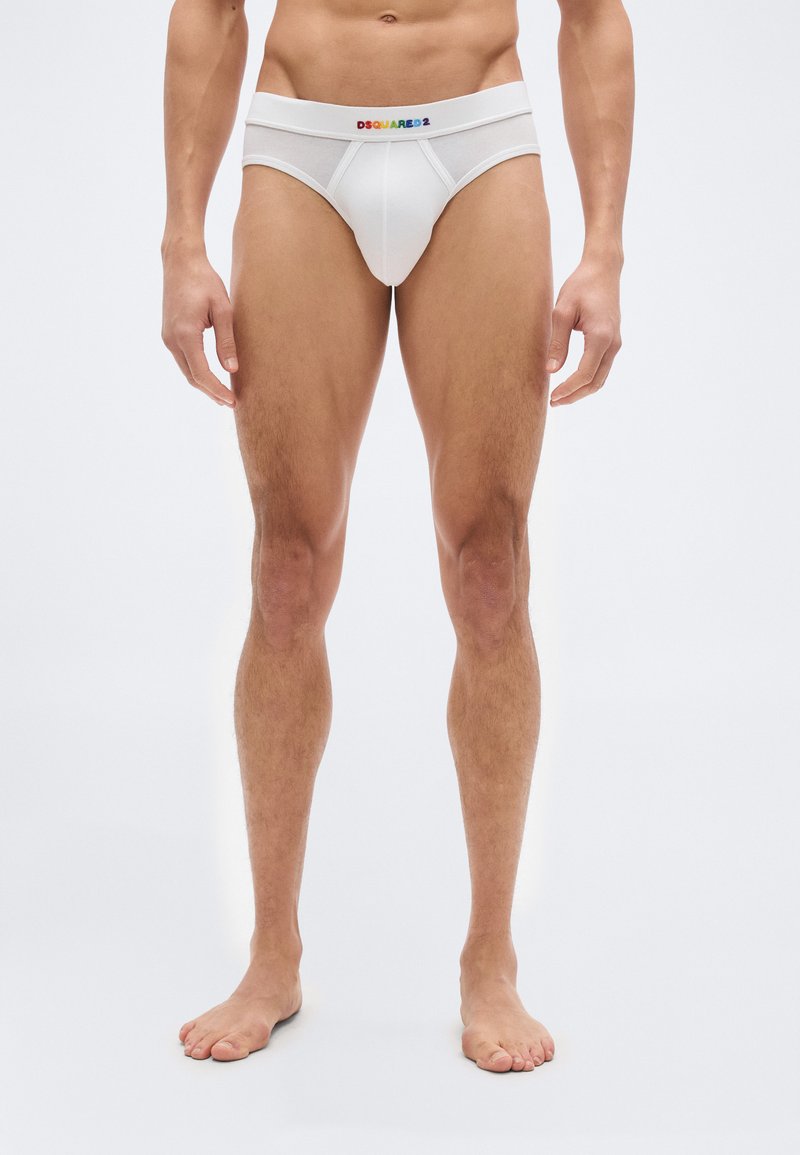 Άνδρας μοντέλο που φοράει λευκά εσώρουχα με πολύχρωμο λογότυπο "DSQUARED2" στη μέση, στέκεται ξυπόλυτος μπροστά σε λευκό φόντο.