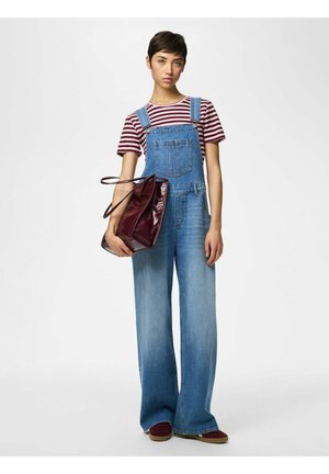 Mladi oseba, oblečena v črtasto majico in široke jeans hlače z naramnicami, drži veliko temno rdečo torbo na belem ozadju.