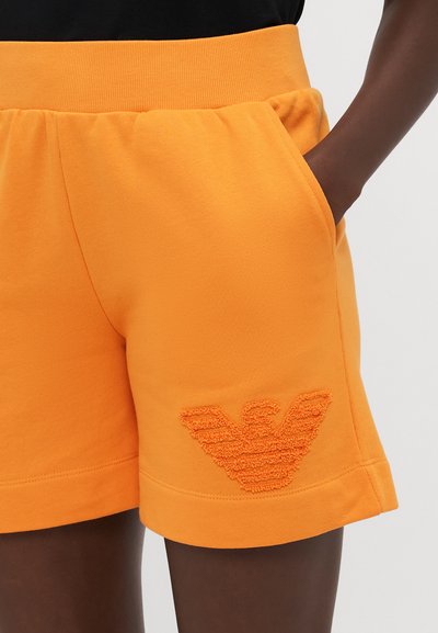Orange cotton shorts with a textured logo patch featuring an emblem on the left thigh. Designed with pockets and an elastic waistband.