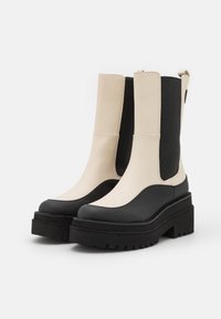 Botas Chelsea de dos tonos con un diseño robusto, con parte superior en color beige, paneles laterales elásticos negros, plataformas negras y suelas con tacos, fotografiadas sobre un fondo blanco.