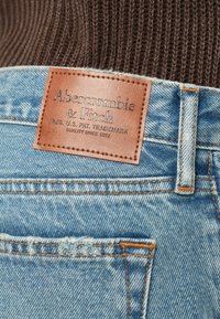 Τζιν τζιν σε ανοιχτό μπλε με δερμάτινο ταμπελάκι με την επιγραφή "Abercrombie & Fitch." Οι ραφές είναι πορτοκαλί και το ύφασμα παρουσιάζει ελαφριά φθορά στις τσέπες.