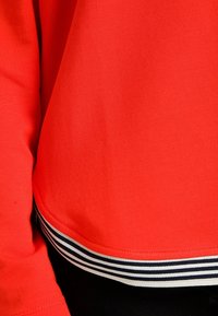 Suéter rojo con una textura suave, que presenta un dobladillo curvado y un acento a rayas en blanco y negro en el borde inferior.