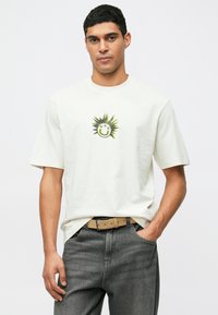 Man som bär en off-white T-shirt med en svart och gul smiley solgrafik och texten "HUGO", grå jeans och beige bälte, med handen i fickan.