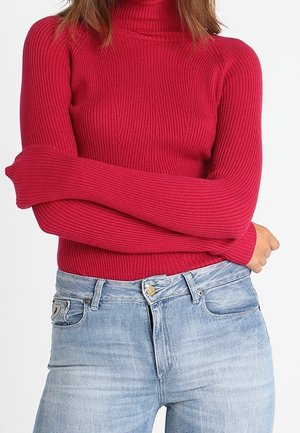 Vrouw draagt een aangesloten rode geribbelde coltrui met lange mouwen, met gekruiste armen over een lichtblauwe spijkerbroek.