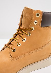 Botín de cuero marr�ón hasta el tobillo con ojetes dorados, cordones amarillos y rojos, y un cuello acolchado en marrón oscuro. Presenta la marca de impermeabilidad en el lateral.