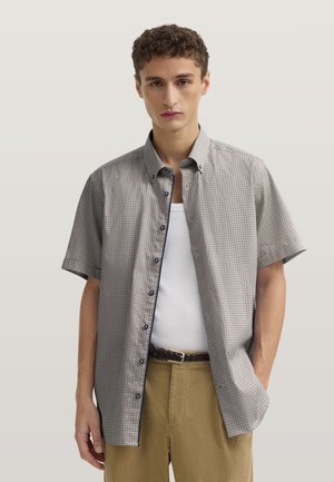 Junger Mann trägt ein kurzärmeliges gemustertes Hemd mit Knöpfen über einem weißen Tanktop, beige Hose und einen geflochtenen braunen Gürtel.