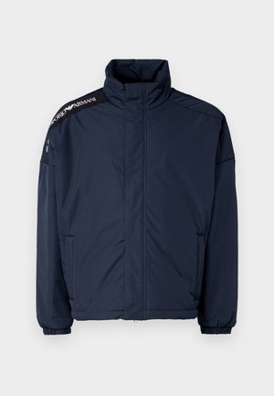 Navy blue puffer jacket with a high neck, elastic cuffs, and zippered pockets. Features black shoulder accents with logo text. Smooth texture.