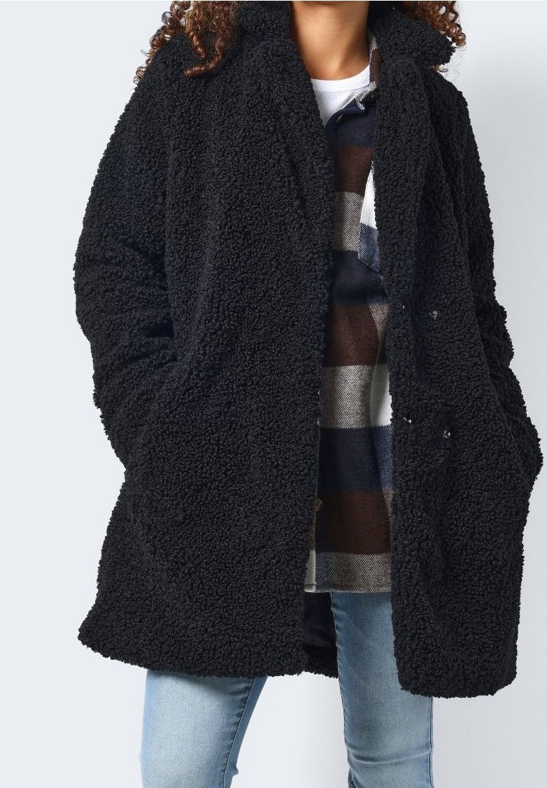 Svart, fluffig, överdimensionerad kappa gjord av texturerat tyg. Har breda slag, två framfickor och en lös passform, lagrad över en rutiga skjorta.