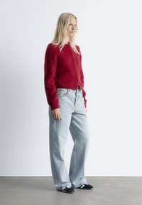 Roter, flauschiger Cardigan mit Knöpfen, kombiniert mit hellblauen, weit geschnittenen Jeans und marineblauen Slippers. Der schlichte Hintergrund lenkt die Aufmerksamkeit auf die Kleidung.