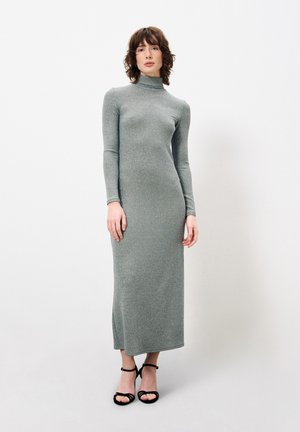 Robe grise à manches longues et col montant avec une coupe ajustée, fabriquée dans un tissu texturé et scintillant ; fente sur le côté à l'ourlet ; associée à des talons aiguilles noirs à brides.